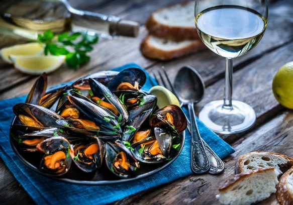 High angle view of steamed mussels in a black plate and white wine glass shot on rustic wooden table. Sliced lemon and bread pieces complete the composition. Selective focus on mussels. Predominant colors are black, orange and blue. XXXL 42Mp studio photo taken with Sony A7rii and Sony FE 90mm f2.8 macro G OSS lens