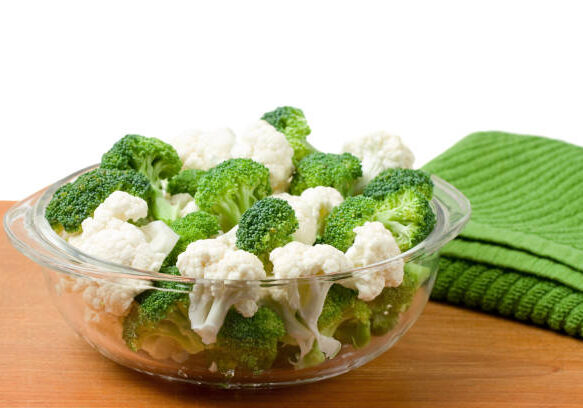 A glass bowl of cauliflower and broccoli on a wood cutting board.